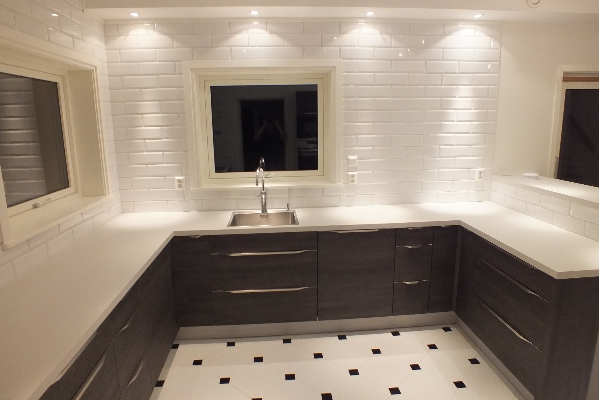 Nowoczesna kuchnia z białymi płytkami typu metro i ciemnymi szafkami. Kontrastowe, czarno-białe płytki podłogowe. Jasne oświetlenie punktowe podkreśla czystość i minimalizm.