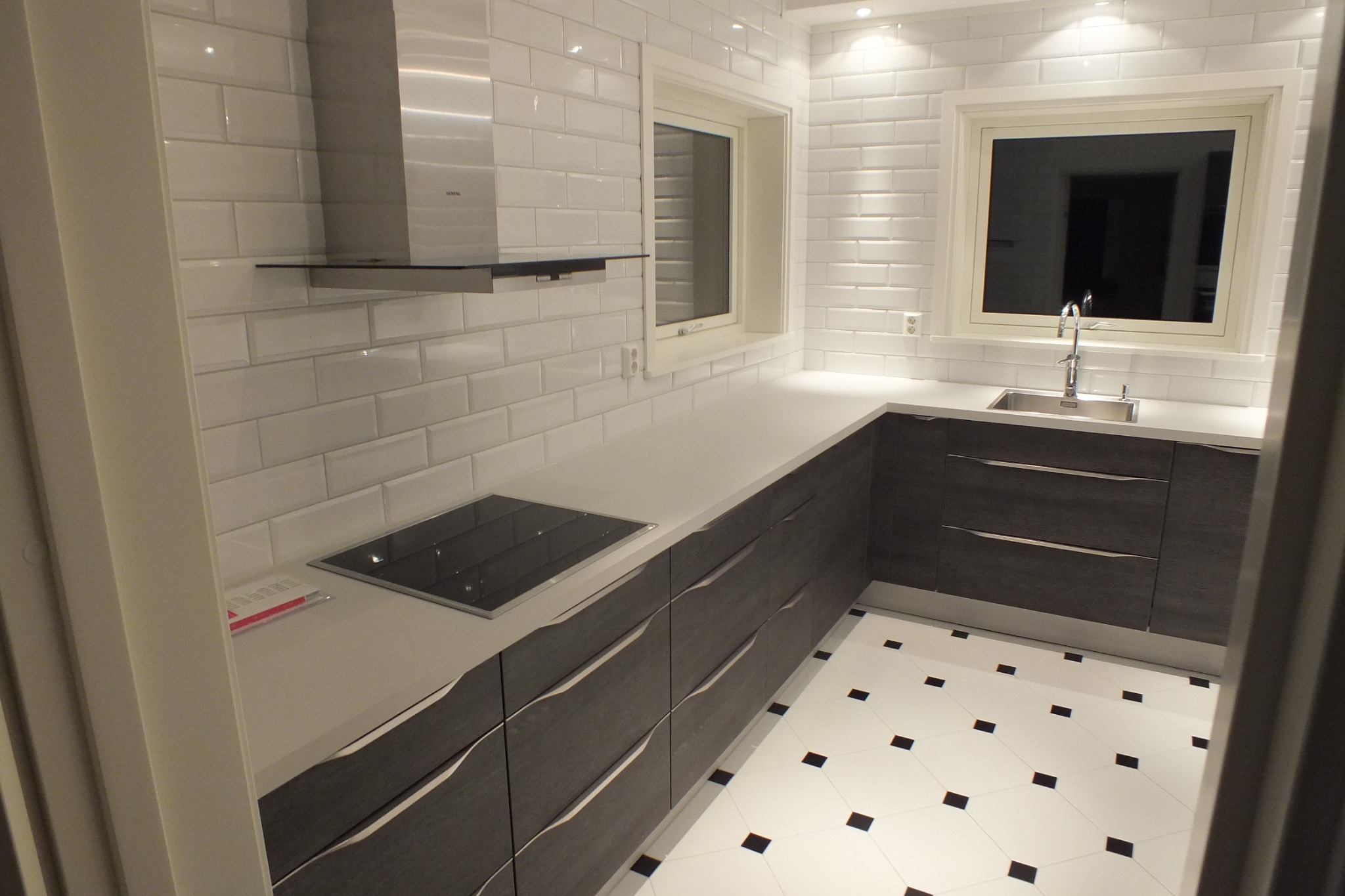 Nowoczesna kuchnia z białymi płytkami, ciemnymi szafkami i białym blatem. Widoczna płyta indukcyjna, okap i okno z widokiem na zewnątrz. Czarno-biała podłoga w karo.