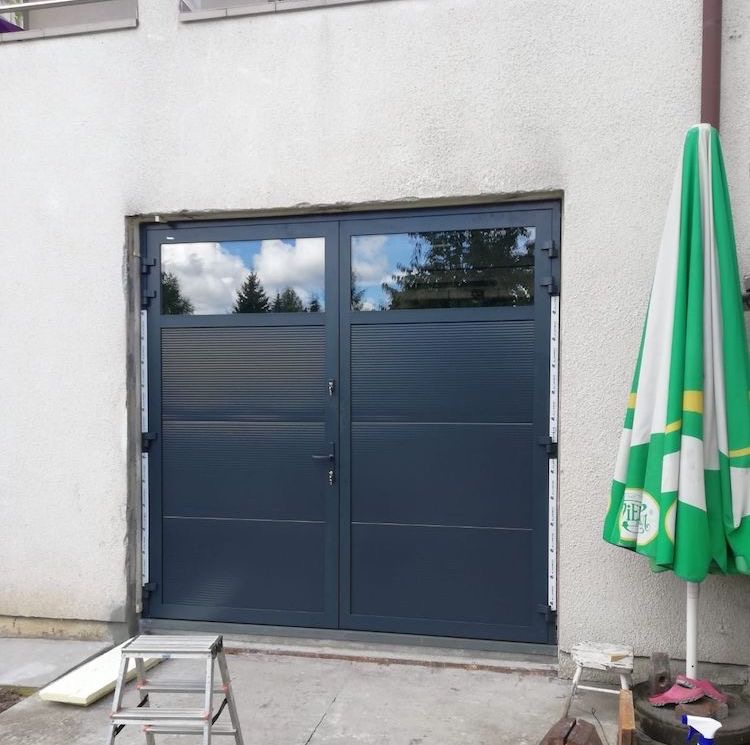 Grafitowe drzwi garażowe z przeszkleniami, odbijające błękitne niebo, na tle białej ściany. Obok rozłożony parasol ogrodowy i drabinka.