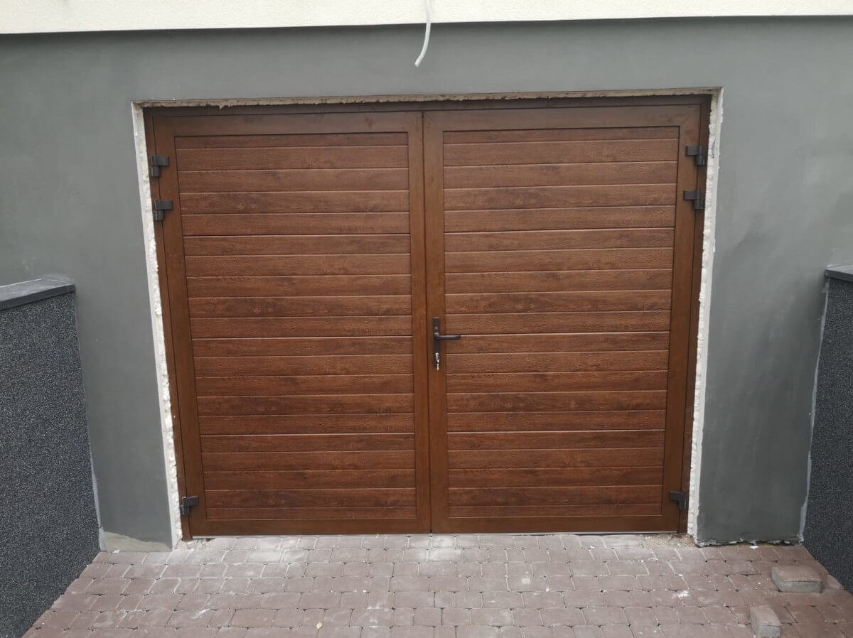 Brązowa, dwuskrzydłowa brama garażowa z poziomymi panelami, zamontowana w szarej ścianie. Widoczna kostka brukowa przed bramą i fragment ściany z boku.