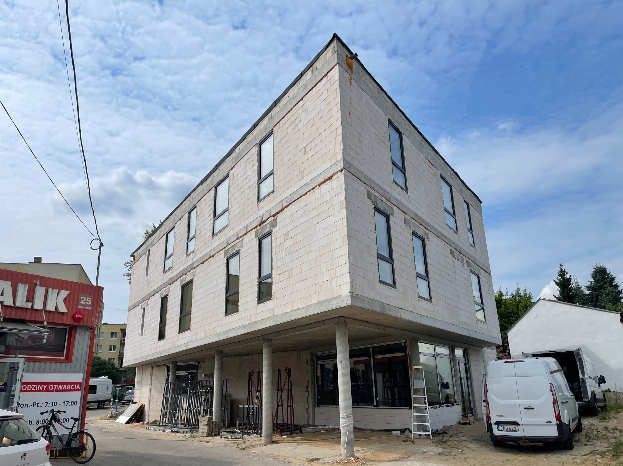 Surowy budynek dwukondygnacyjny z oknami, w trakcie budowy, z widocznymi elementami konstrukcyjnymi i rusztowaniami. Biały van dostawczy w tle.