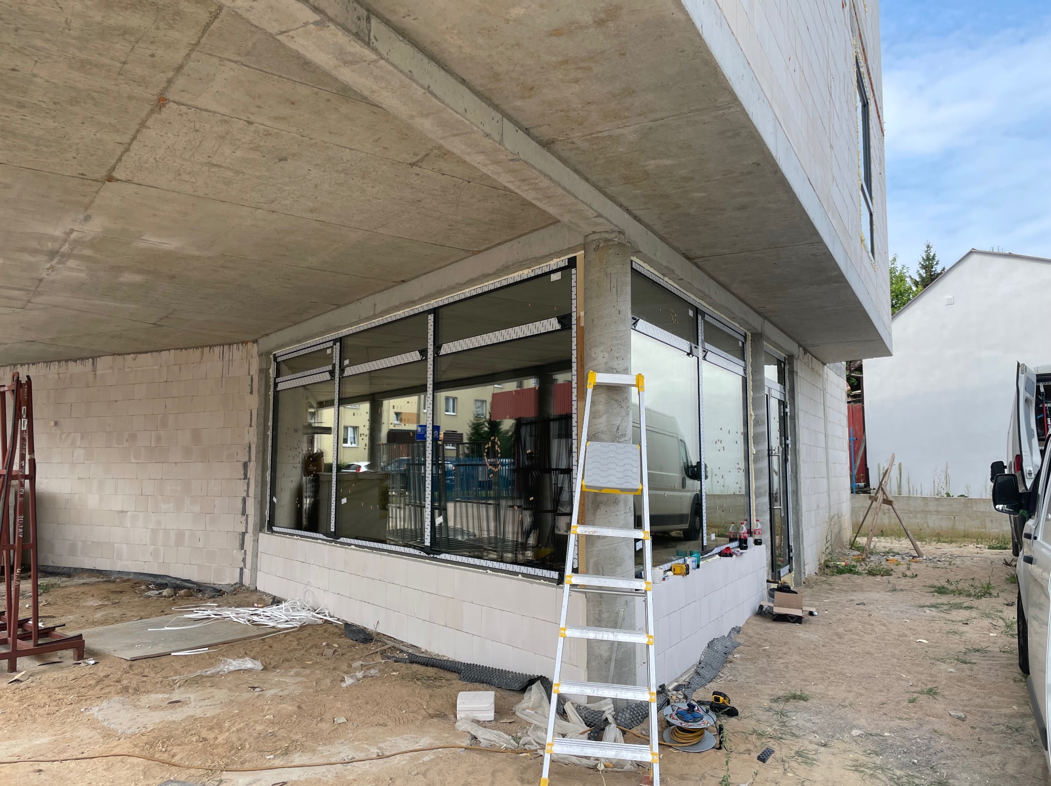 Montaż okien w surowym budynku z betonu komórkowego. Widoczna drabina, narzędzia i częściowo zamontowane okna z czarnymi ramami. W tle zaparkowany biały bus.