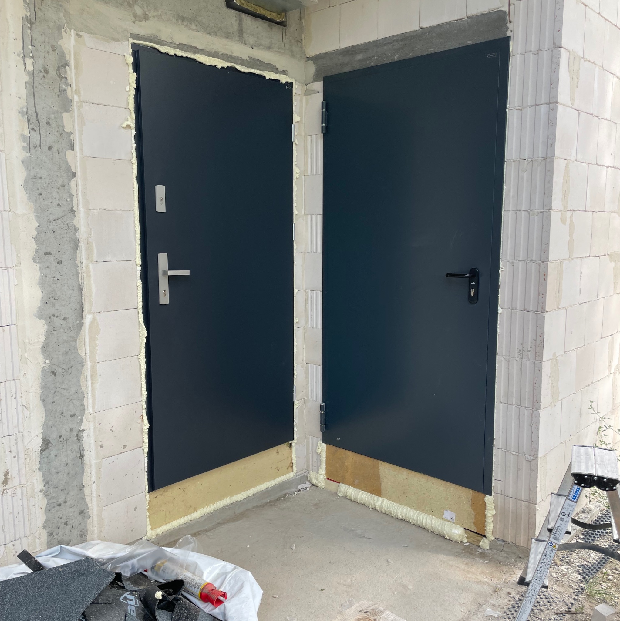 Montaż dwóch grafitowych drzwi zewnętrznych w surowym stanie. Widoczna pianka montażowa i fragment drabiny. Drzwi osadzone w murze z betonu komórkowego.