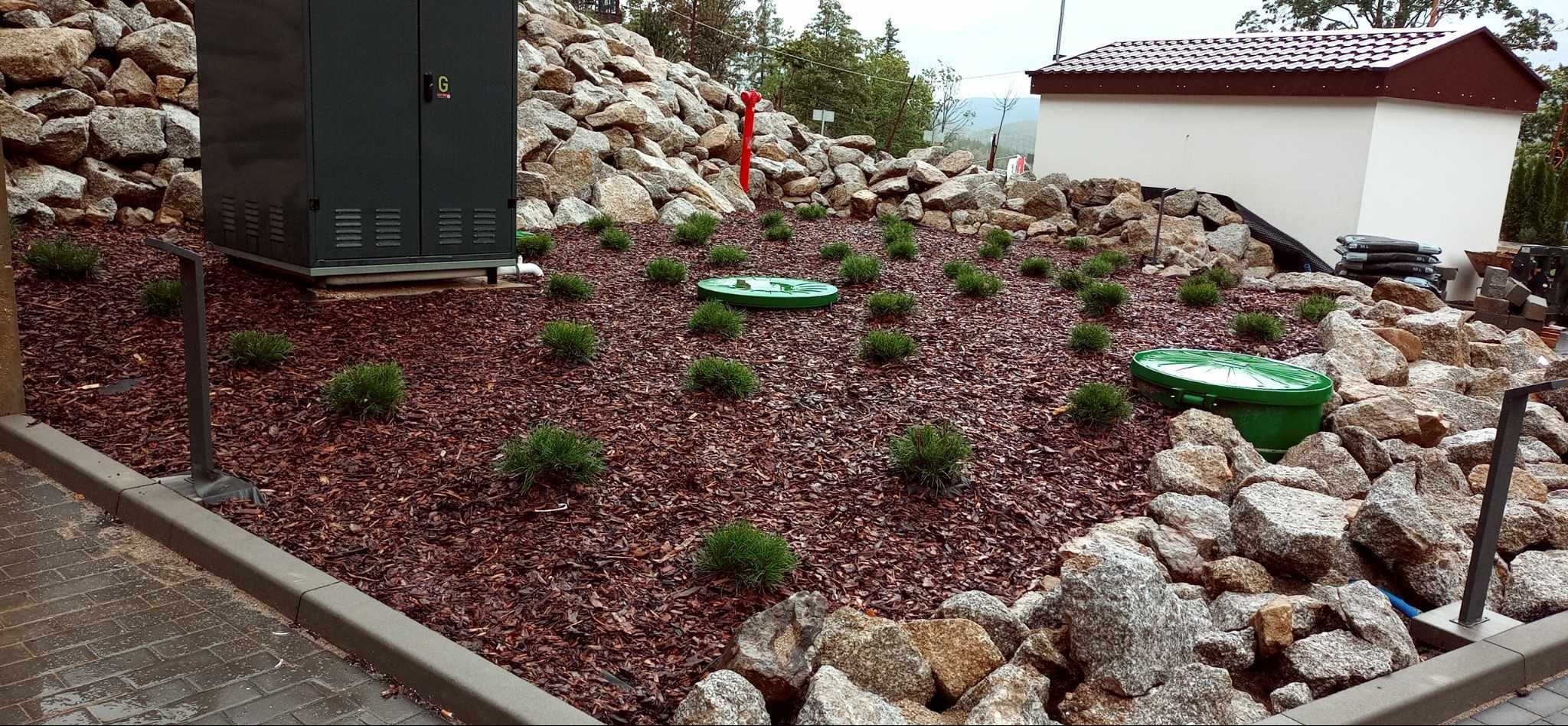 Aranżacja ogrodu: rabata z kory, niskie krzewy iglaste, kamienie, zielone pokrywy studzienek. W tle transformator i budynek.