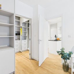 NIEBANALNA BROKERKA SPÓŁKA Z OGRANICZONĄ ODPOWIEDZIALNOŚCIĄ - Jasne wnętrze mieszkania z garderobą i widokiem na kuchnię. Drewniana podłoga, białe ściany i meble. Wazon z rośliną i lampka na stoliku nocnym.