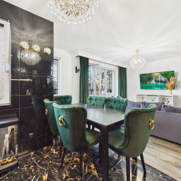 NIEBANALNA BROKERKA - Elegancka jadalnia z czarnym marmurem, zielonymi krzesłami i kryształowymi żyrandolami. Widok na salon z szarą sofą i telewizorem. Nowoczesne wykończenie wnętrz.