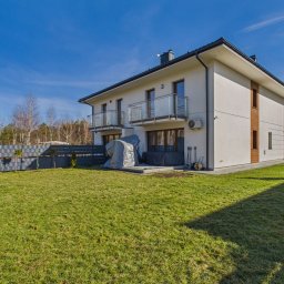 NIEBANALNA BROKERKA SPÓŁKA Z OGRANICZONĄ ODPOWIEDZIALNOŚCIĄ - Dwukondygnacyjny dom z balkonami i zadbanym trawnikiem w słoneczny dzień. Widoczny fragment ogrodzenia i zaparkowany samochód. Dom w stylu nowoczesnym.
