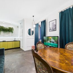 NIEBANALNA BROKERKA SPÓŁKA Z OGRANICZONĄ ODPOWIEDZIALNOŚCIĄ - Jasne wnętrze: salon z drewnianym stołem, zieloną sofą i kuchnią w tle. Białe ściany, parkiet w jodełkę i turkusowe zasłony dodają elegancji.