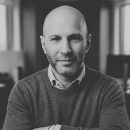 NIEBANALNA BROKERKA SPÓŁKA Z OGRANICZONĄ ODPOWIEDZIALNOŚCIĄ - Czarno-biały portret mężczyzny z brodą, w swetrze, z założonymi rękami, patrzącego wprost, z oknem w tle. Profesjonalny wizerunek agenta nieruchomości.