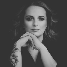 NIEBANALNA BROKERKA SPÓŁKA Z OGRANICZONĄ ODPOWIEDZIALNOŚCIĄ - Portret kobiety z tatuażem na ramieniu, w czerni i bieli, z rękami pod brodą, patrzącej wprost, z delikatnym makijażem i kolczykami.