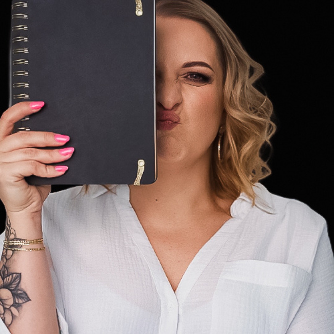 Kobieta z blond lokami i różowymi paznokciami, trzymająca czarny notatnik, zasłaniający część twarzy, w białej koszuli i złotych kolczykach.