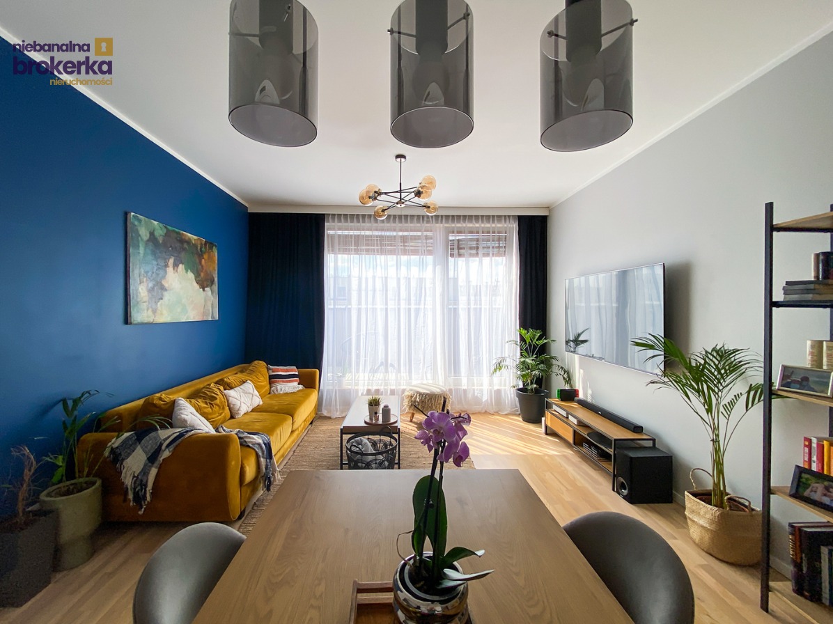 Nowoczesny salon z żółtą sofą, niebieską ścianą i drewnianym stołem. Widoczne trzy szare lampy wiszące i duży telewizor. Jasne, przestronne wnętrze z roślinami.