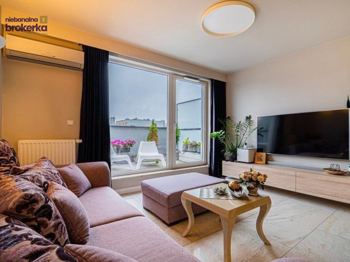 Jasny salon z sofą w odcieniach różu, balkonem z widokiem na miasto i telewizorem. Nowoczesne wnętrze z elementami dekoracyjnymi i kwiatami.