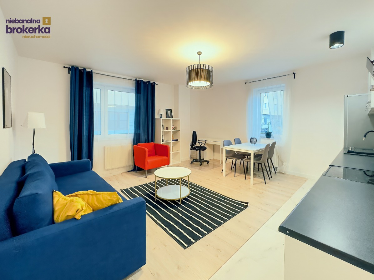 Jasne wnętrze mieszkania: niebieska sofa z żółtymi poduszkami, okrągły stolik na dywanie w paski, czerwony fotel i stół z krzesłami w tle. Widok na okno i elementy kuchni.