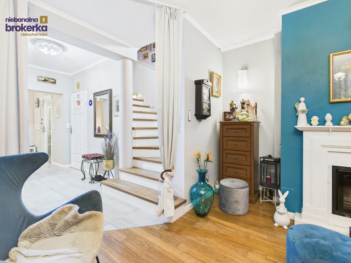 Jasne wnętrze domu z drewnianymi schodami i elementami dekoracyjnymi: komoda, zegar, wazon z kwiatami, fotel. Ściana w odcieniach błękitu z kominkiem.