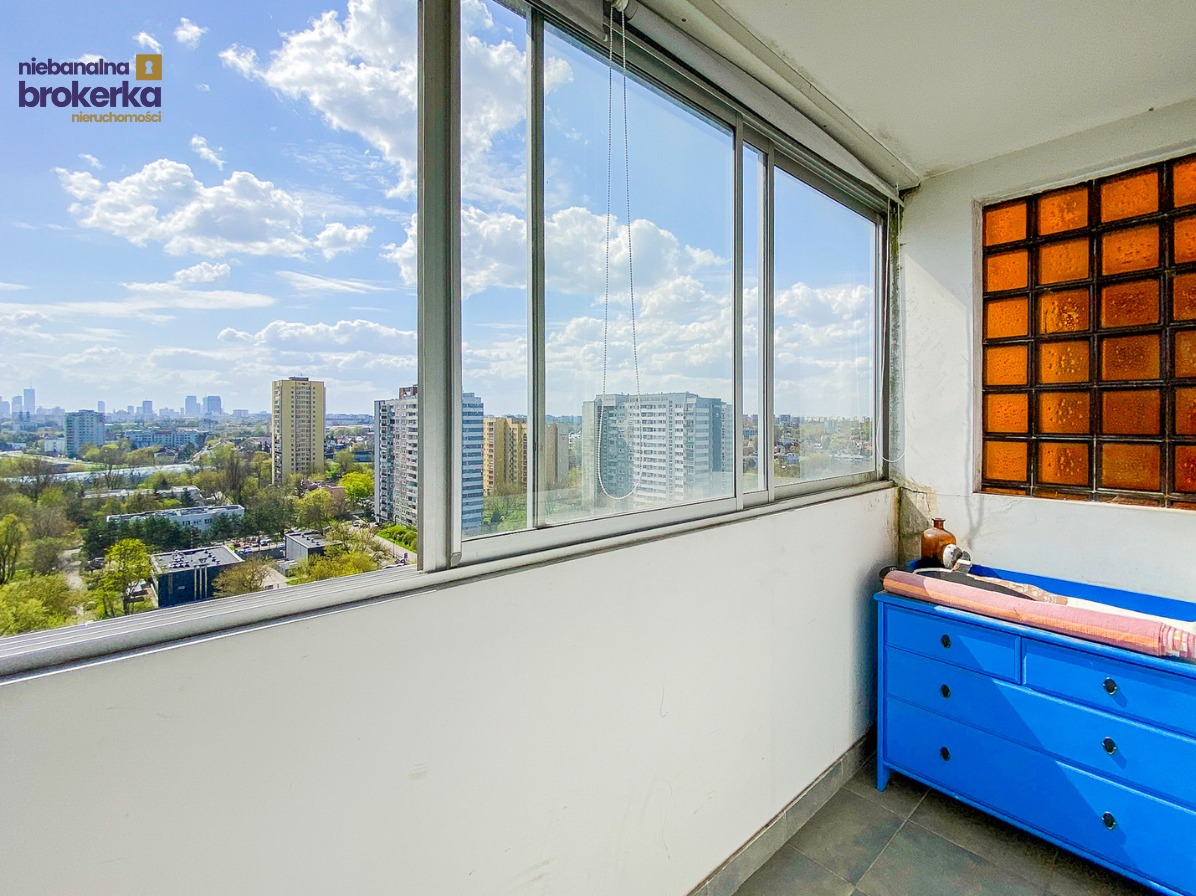 Loggia z widokiem na panoramę miasta przez duże okna. Wnętrze zdobi błękitna komoda i pomarańczowe luksfery. Słoneczny dzień, błękitne niebo z chmurami.