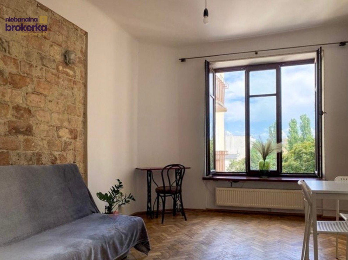 Jasne, przestronne wnętrze mieszkania w Warszawie z ceglaną ścianą, dużym oknem i parkietem. Widok na zieleń za oknem.