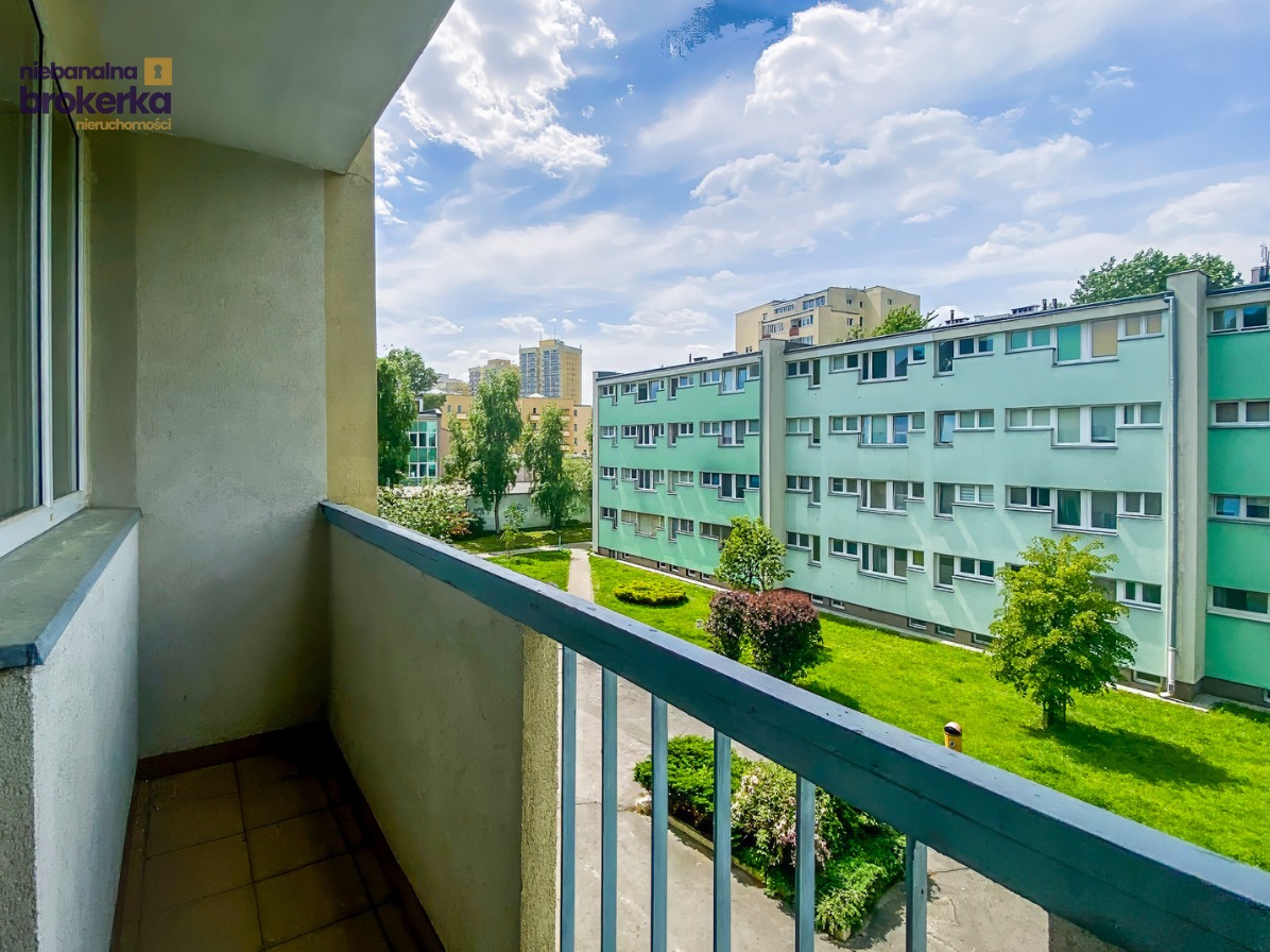 Widok z balkonu na zielony dziedziniec i budynek mieszkalny w słoneczny dzień. Balustrada balkonu w kolorze szarym. W tle błękitne niebo z chmurami.