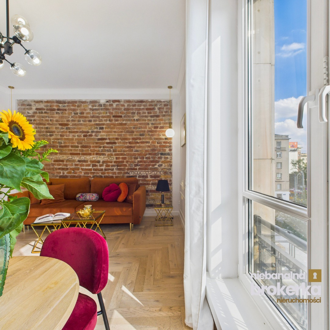 Stylowe wnętrze: ceglana ściana, pomarańczowa sofa z poduszkami, parkiet w jodełkę i bukiet słoneczników, widok z okna na miasto. Wystrój z charakterem.