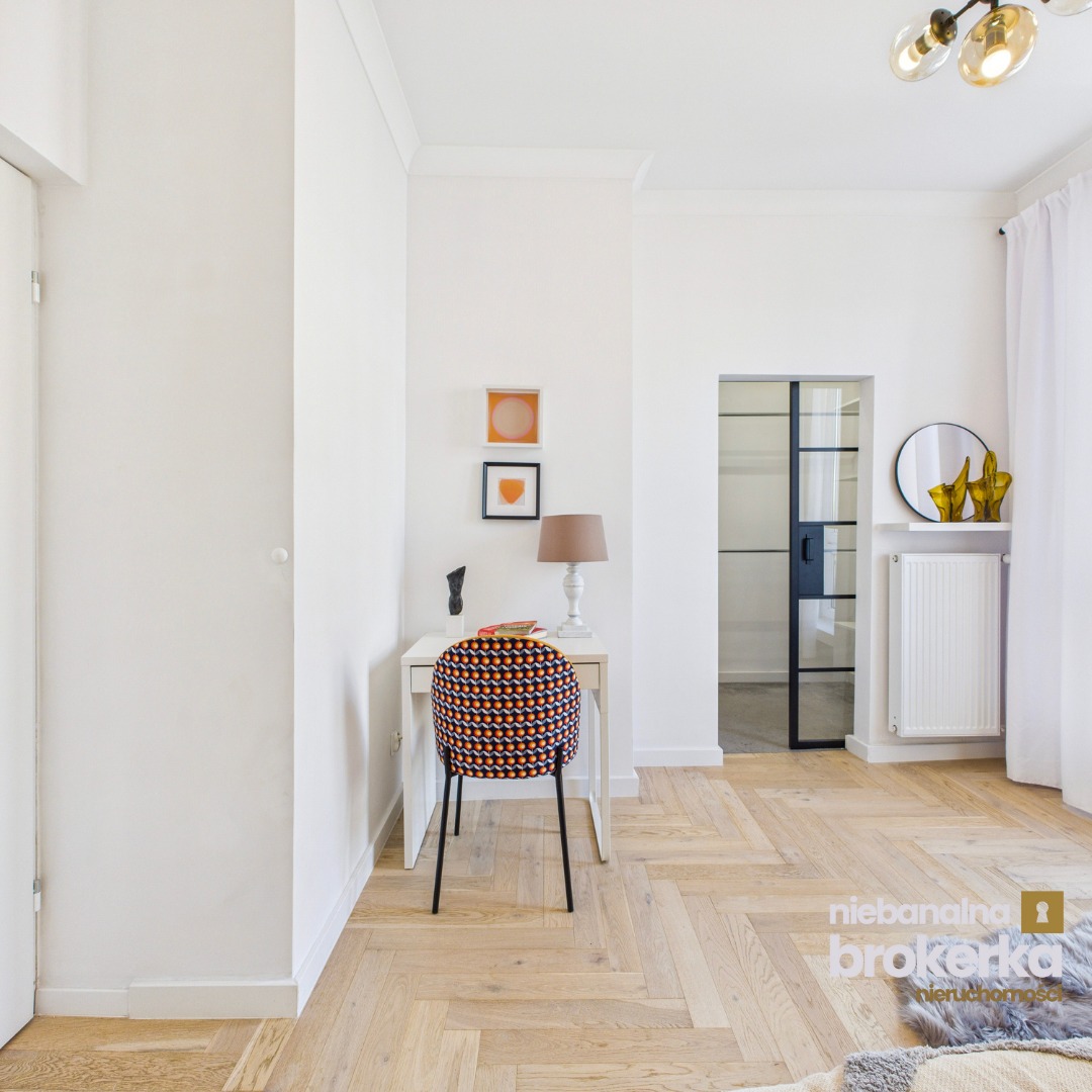 Jasne wnętrze z parkietem w jodełkę, biurkiem, krzesłem w geometryczny wzór i czarną, przeszkloną ścianką działową. Nowoczesny design z akcentami retro.