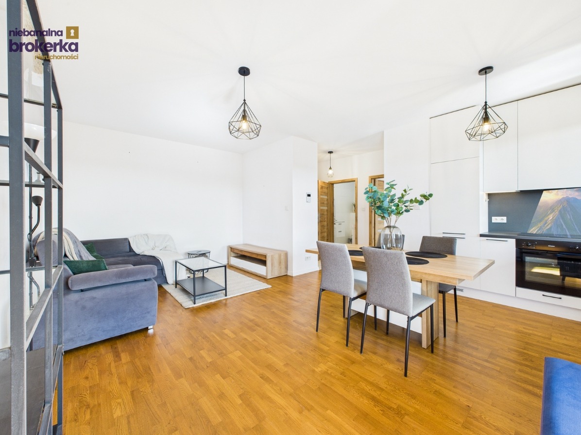 Jasne wnętrze mieszkania z drewnianą podłogą, szarą sofą, stołem jadalnym i nowoczesną kuchnią. Widoczne dekoracje i oświetlenie.