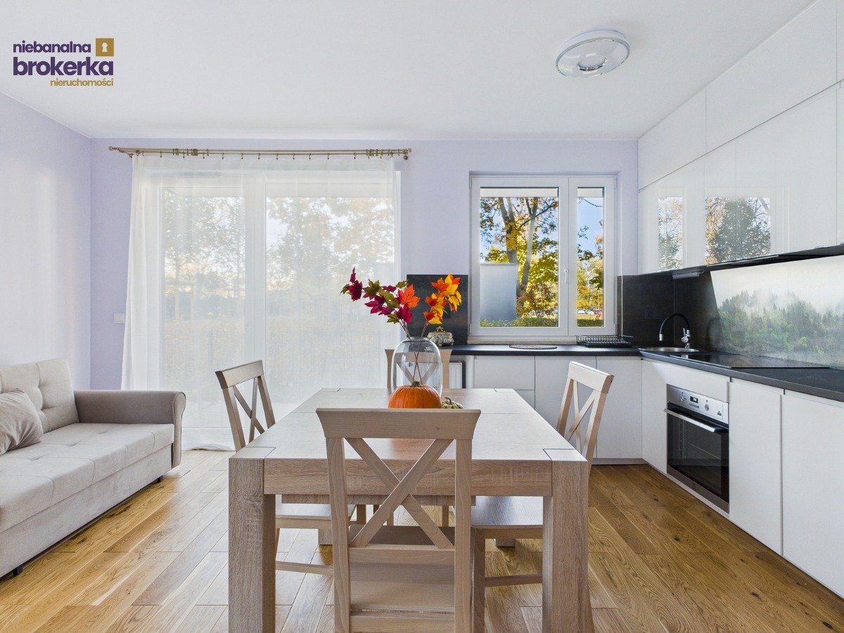 Jasne wnętrze: stół z dynią i jesiennymi liśćmi, sofa, biała kuchnia z leśnym motywem na ścianie. Widok z okna na drzewa w jesiennej scenerii.