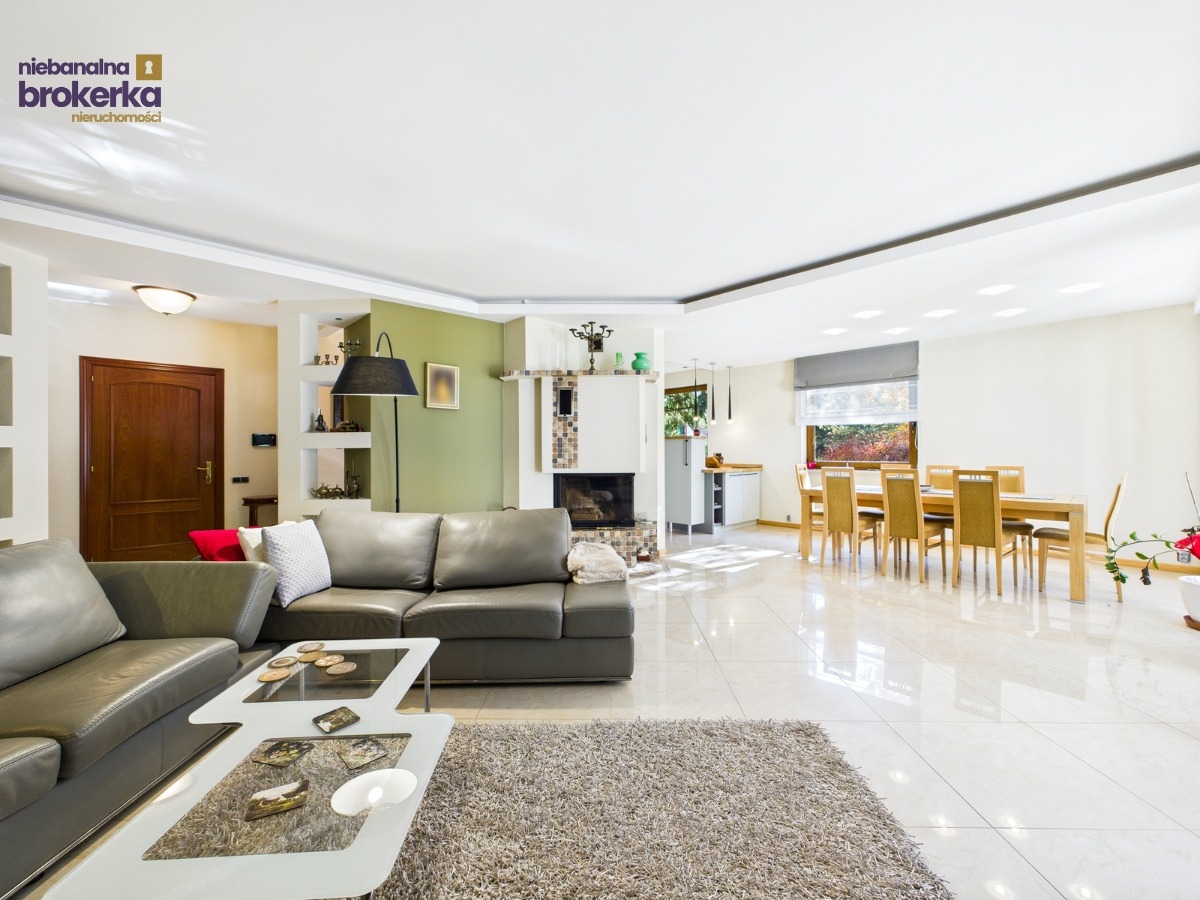 Jasne, przestronne wnętrze z szarymi sofami, nowoczesnym stolikiem kawowym i kominkiem. Widoczna jadalnia z oknem. Marmurowe podłogi odbijają światło.