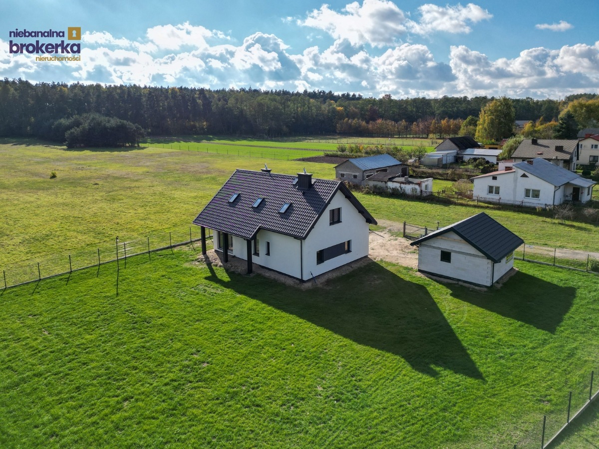 Nowoczesny dom z ciemnym dachem i jasną elewacją, otoczony zielonym trawnikiem i ogrodzeniem. Widok z lotu ptaka, w tle las i zabudowania.