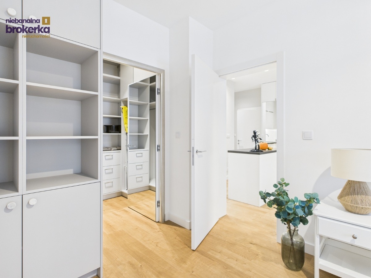 Jasne wnętrze mieszkania z garderobą i widokiem na kuchnię. Drewniana podłoga, białe ściany i meble. Wazon z rośliną i lampka na stoliku nocnym.