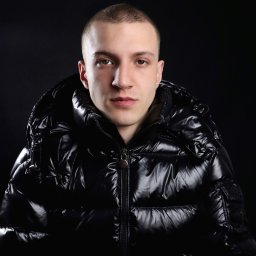 Martyna Kucharska - Mężczyzna w czarnej, błyszczącej kurtce puchowej z kapturem na ciemnym tle. Portret z przodu, oświetlenie studyjne podkreśla detale kurtki.