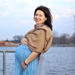 Martyna Kucharska - Uśmiechnięta kobieta w ciąży, w błękitnej sukience i beżowym szalu, opiera się o barierkę z widokiem na wodę i budynki w tle. Delikatne światło podkreśla naturalność.