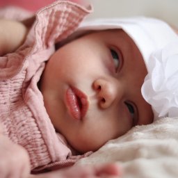 Martyna Kucharska - Portret niemowlęcia w różowej sukience i białym czepku z kwiatem, leżącego na miękkim, jasnym kocyku. Delikatne światło podkreśla rysy twarzy dziecka.