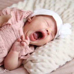 Martyna Kucharska - Noworodek w różowej sukience i białym czepku z kwiatem, leżący na kremowym kocyku, z szeroko otwartymi ustami podczas ziewania. Delikatne, naturalne światło.