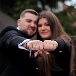 Martyna Kucharska - Uśmiechnięta para prezentuje obrączki ślubne, zbliżając zaciśnięte pięści do obiektywu. Mężczyzna w czarnym garniturze, kobieta w czarnej marynarce. Tło rozmyte.