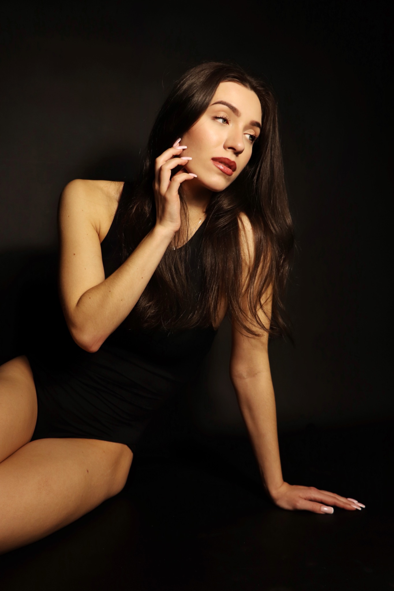 Kobieta w czarnym body pozuje na ciemnym tle, z dłonią przy twarzy i długimi, ciemnymi włosami. Uwagę przyciągają starannie wykonane paznokcie.