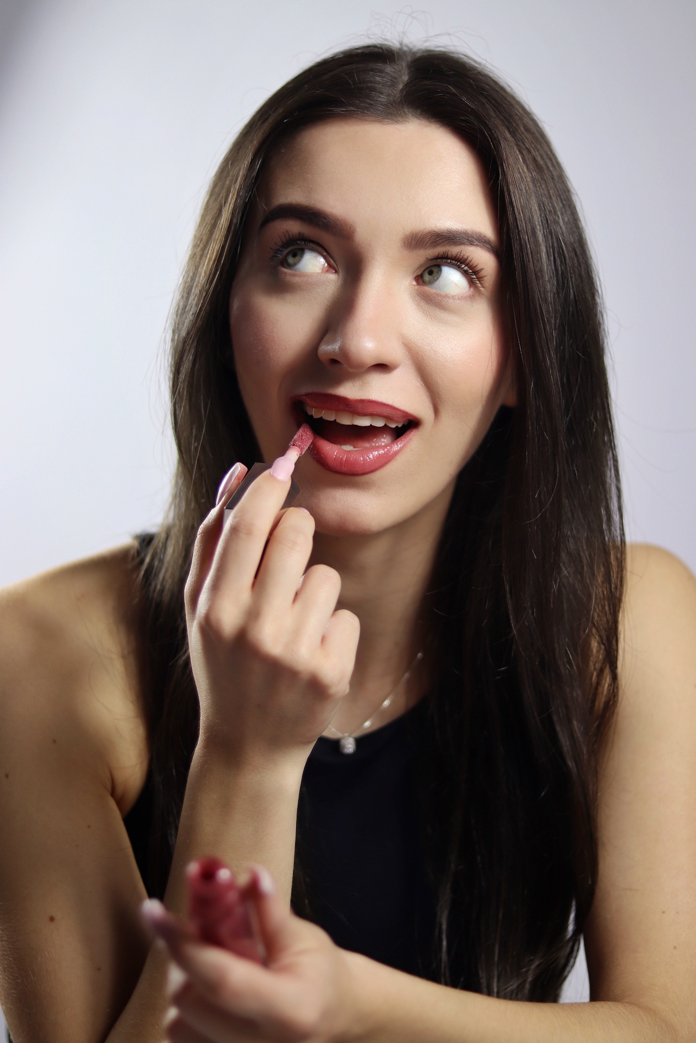 Portret kobiety z długimi, ciemnymi włosami, aplikującej błyszczyk na usta. Uśmiech, spojrzenie w górę, delikatny makijaż, subtelna biżuteria. Jasne tło.