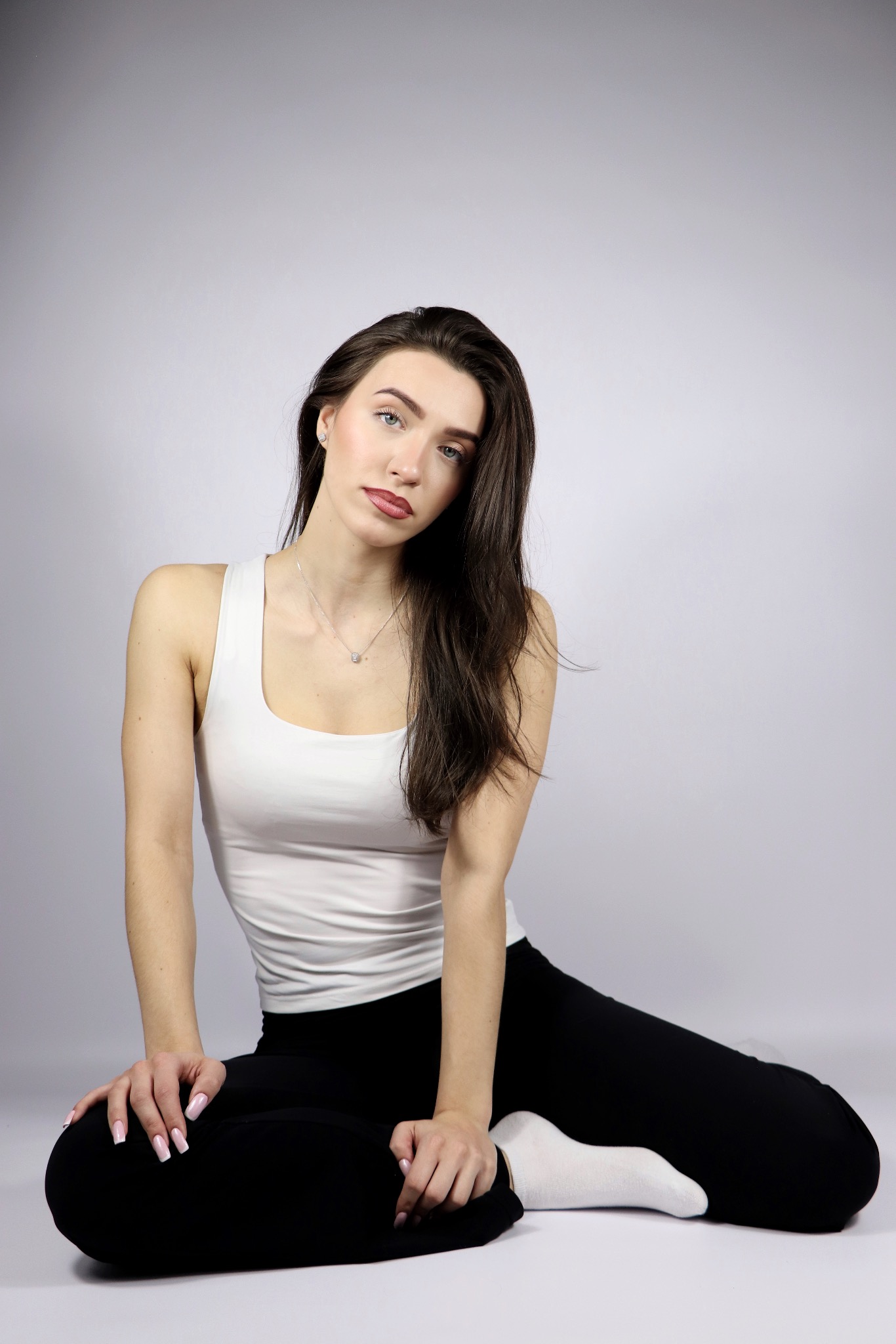 Portret kobiety siedzącej na podłodze w białym topie i czarnych legginsach, z delikatnym makijażem i srebrnym naszyjnikiem na tle jasnoszarego studia.