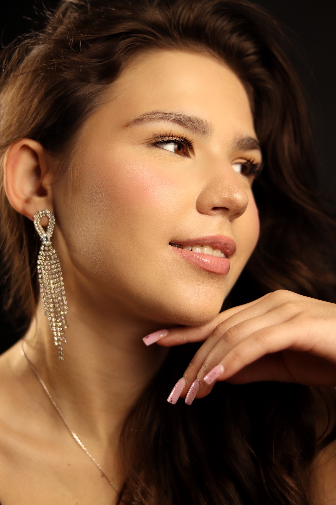 Portret młodej kobiety z błyszczącymi kolczykami i delikatnym makijażem, spoglądającej w prawo, z dłonią pod brodą. Ciemne tło podkreśla urodę modelki.