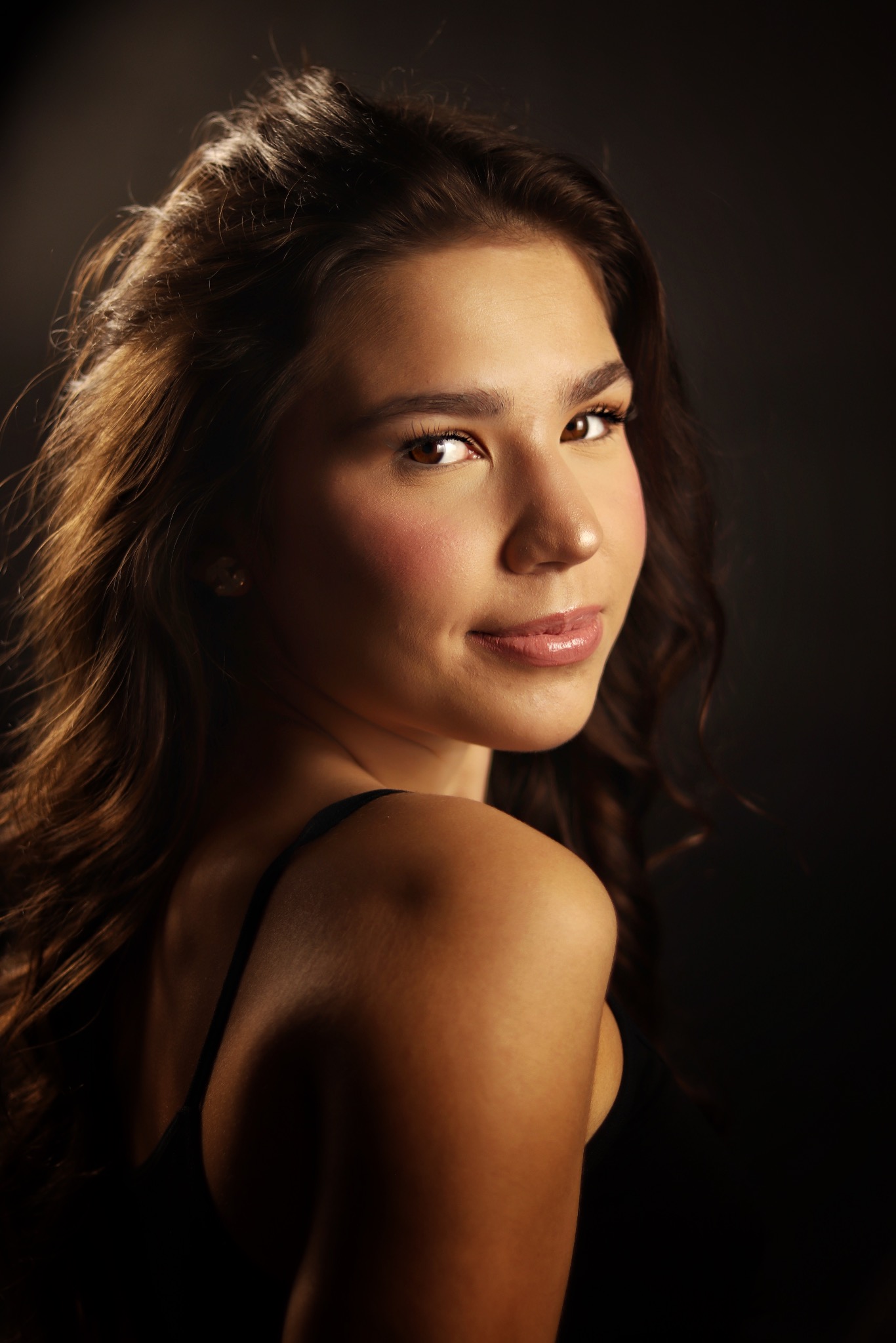 Portret młodej kobiety z długimi, brązowymi włosami, spoglądającej przez ramię, z delikatnym makijażem i ciepłym oświetleniem na ciemnym tle.