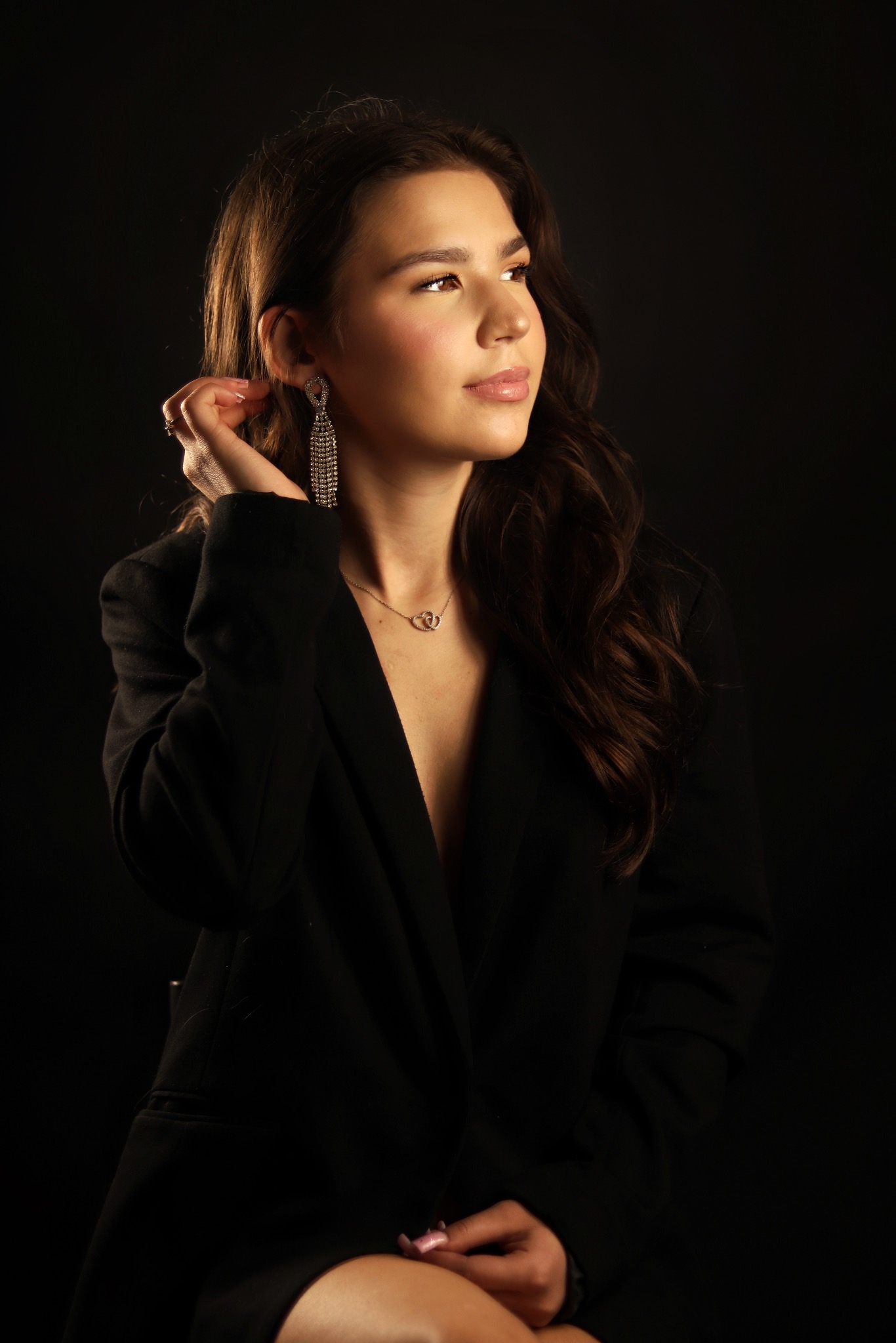 Portret kobiety w czarnej marynarce, z błyszczącymi kolczykami i naszyjnikiem na ciemnym tle. Ujęcie z profilu, delikatny makijaż, profesjonalna sesja.