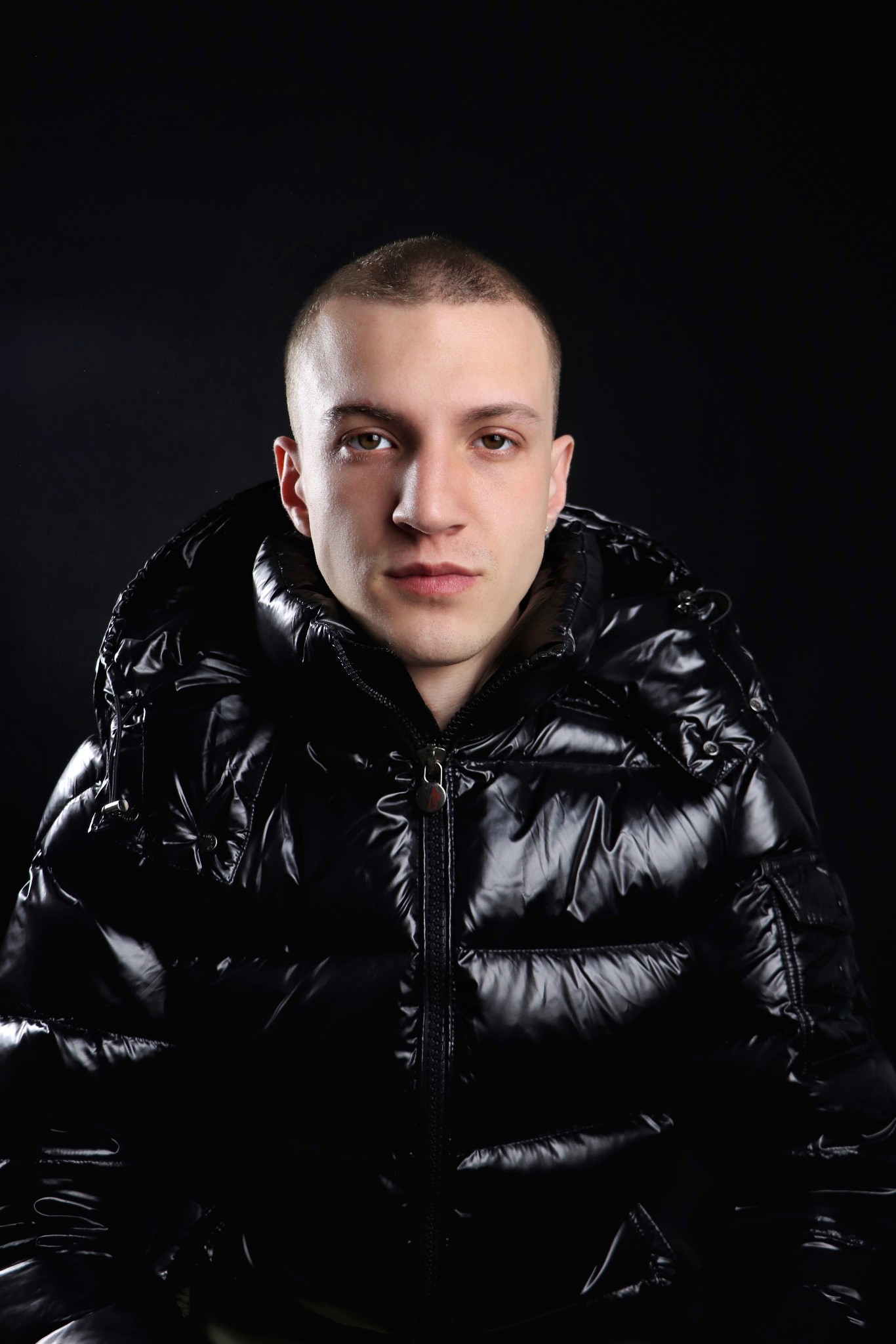 Mężczyzna w czarnej, błyszczącej kurtce puchowej z kapturem na ciemnym tle. Portret z przodu, oświetlenie studyjne podkreśla detale kurtki.