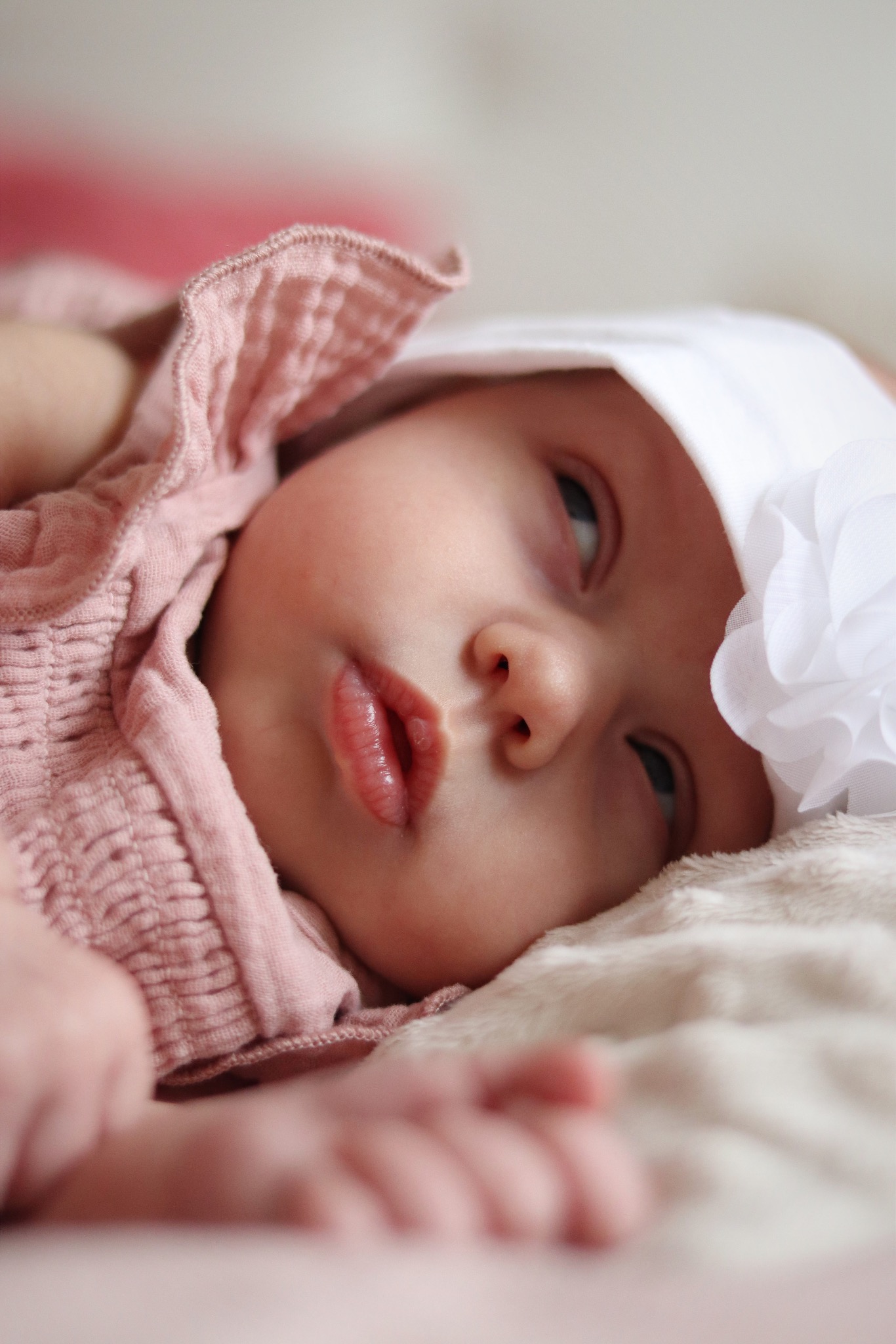 Portret niemowlęcia w różowej sukience i białym czepku z kwiatem, leżącego na miękkim, jasnym kocyku. Delikatne światło podkreśla rysy twarzy dziecka.