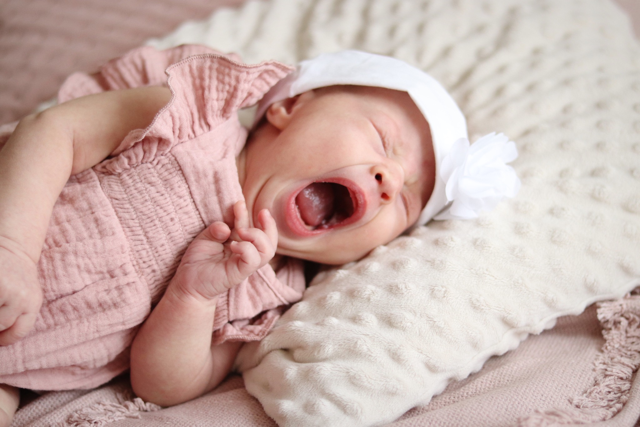 Noworodek w różowej sukience i białym czepku z kwiatem, leżący na kremowym kocyku, z szeroko otwartymi ustami podczas ziewania. Delikatne, naturalne światło.