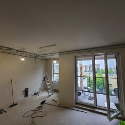 Daniel Dobosz - Wnętrze w trakcie remontu: widoczne profile sufitowe, drabina, narzędzia. Otwarty balkon z widokiem na miasto. Przygotowanie do montażu płyty karton-gips.