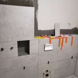 Daniel Dobosz - Montaż płytek w łazience, widoczne narzędzia i akcesoria glazurnicze, otwory na armaturę, jasne płytki z delikatnym wzorem. Etap prac wykończeniowych.