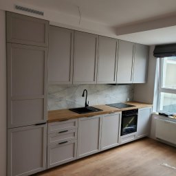 Daniel Dobosz - Jasna kuchnia z szarymi szafkami, drewnianym blatem i marmurowym backsplashiem. Czarny zlew i płyta grzewcza. Okno z roletą rzymską. Nowoczesny design.