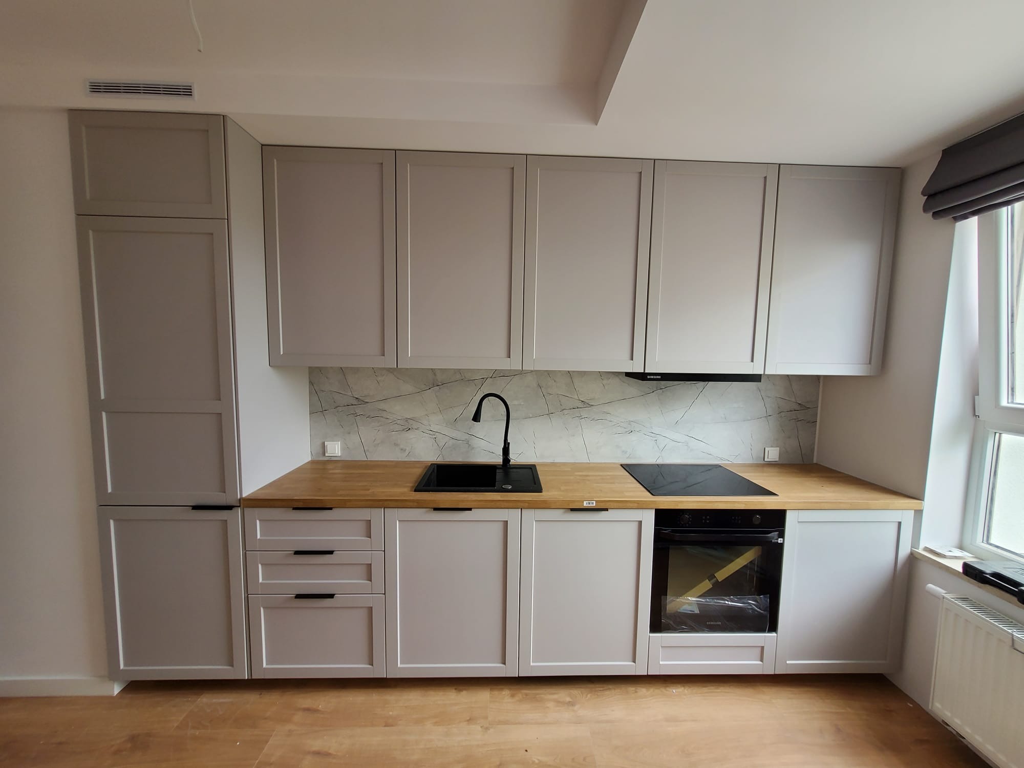 Jasnoszara zabudowa kuchenna z drewnianym blatem i marmurowym panelem nad blatem. Widoczny zlew, płyta indukcyjna i piekarnik. Minimalistyczny styl.