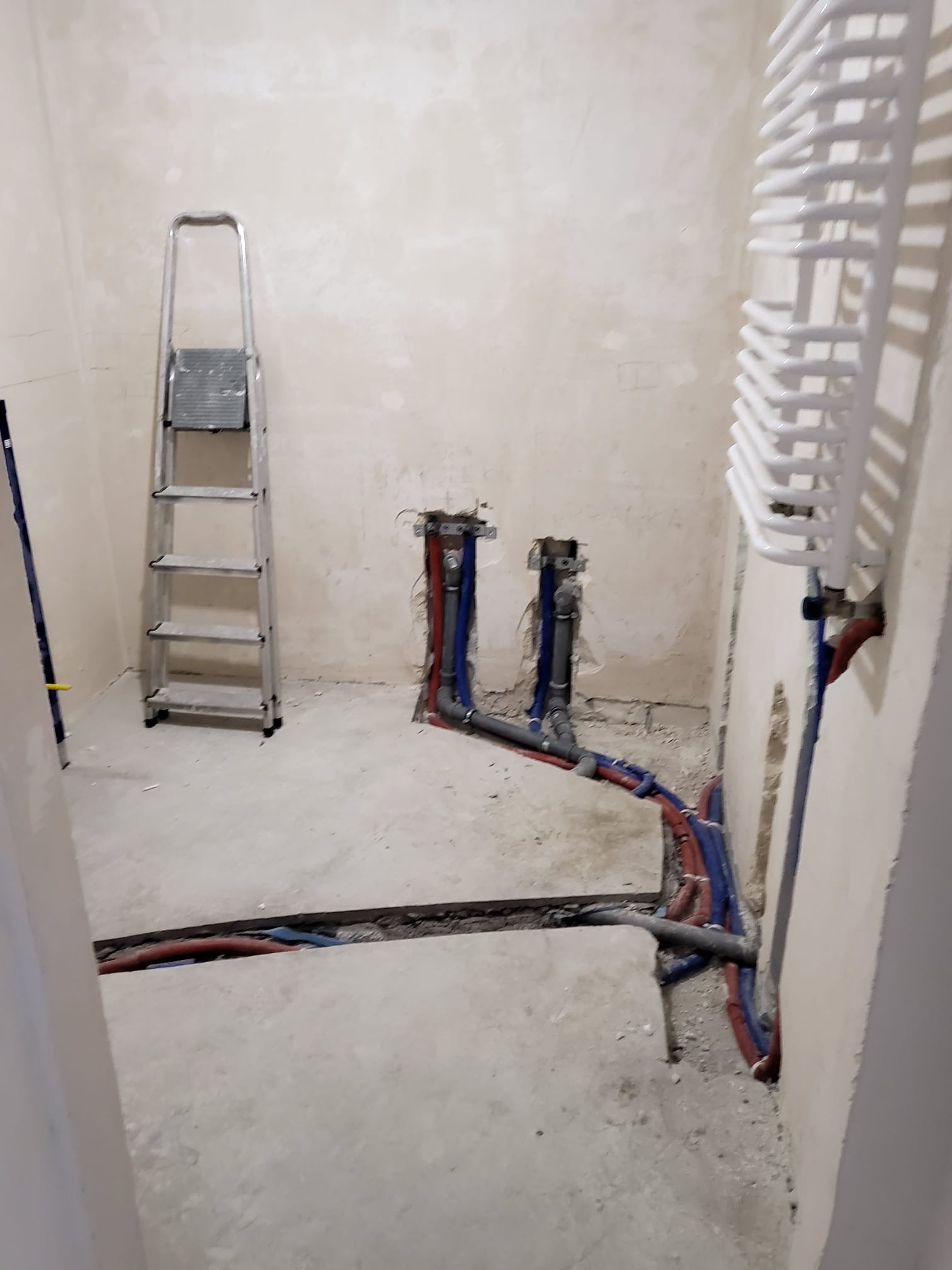 Łazienka w trakcie remontu: widoczne rury instalacji wodnej i grzewczej w ścianach i podłodze. Grzejnik drabinkowy zamontowany. Aluminiowa drabina oparta o ścianę.