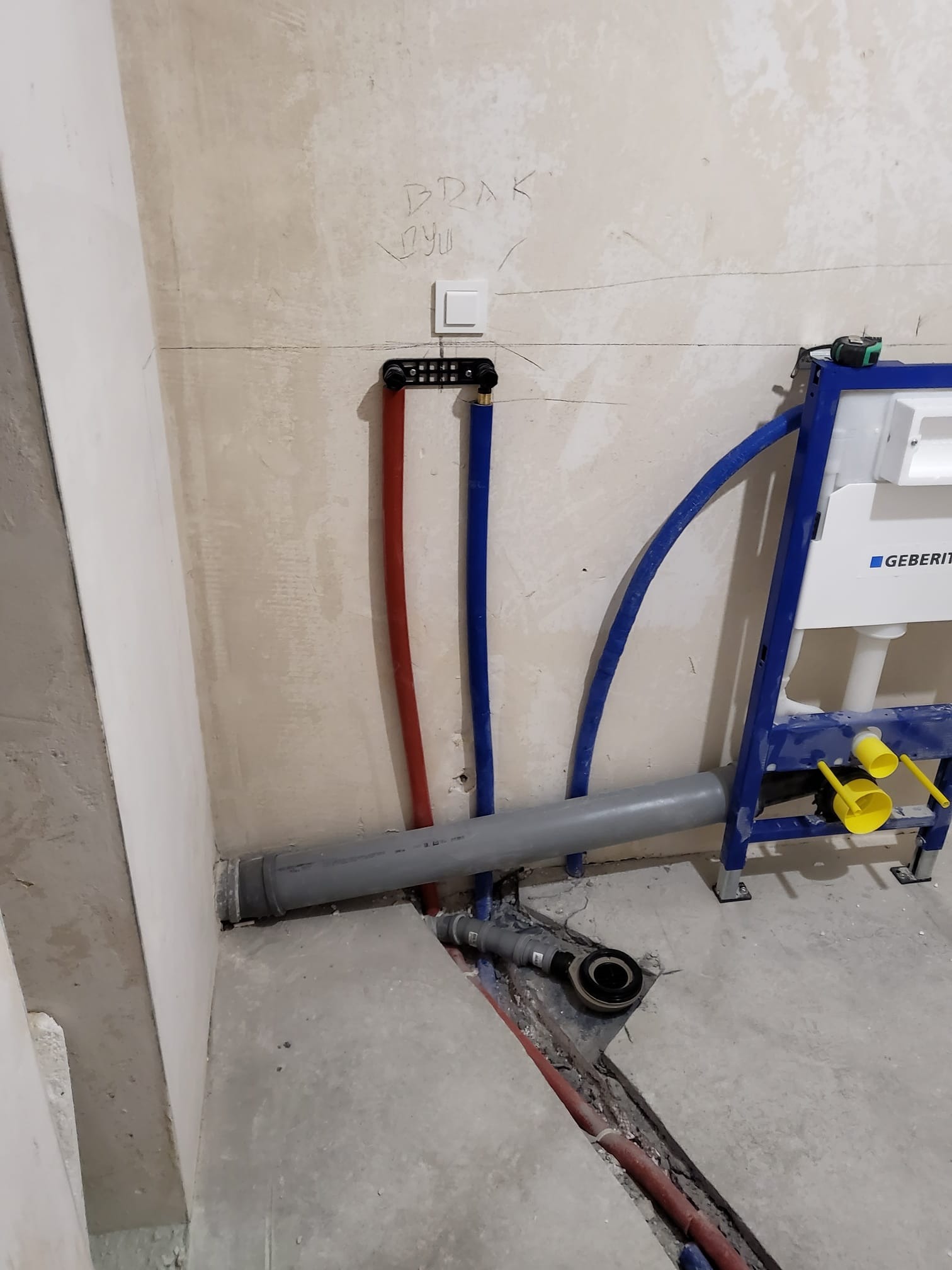 Montaż stelaża podtynkowego Geberit z widocznymi rurami doprowadzającymi wodę. Surowy stan, ściana z napisem. Instalacja sanitarna w trakcie prac.