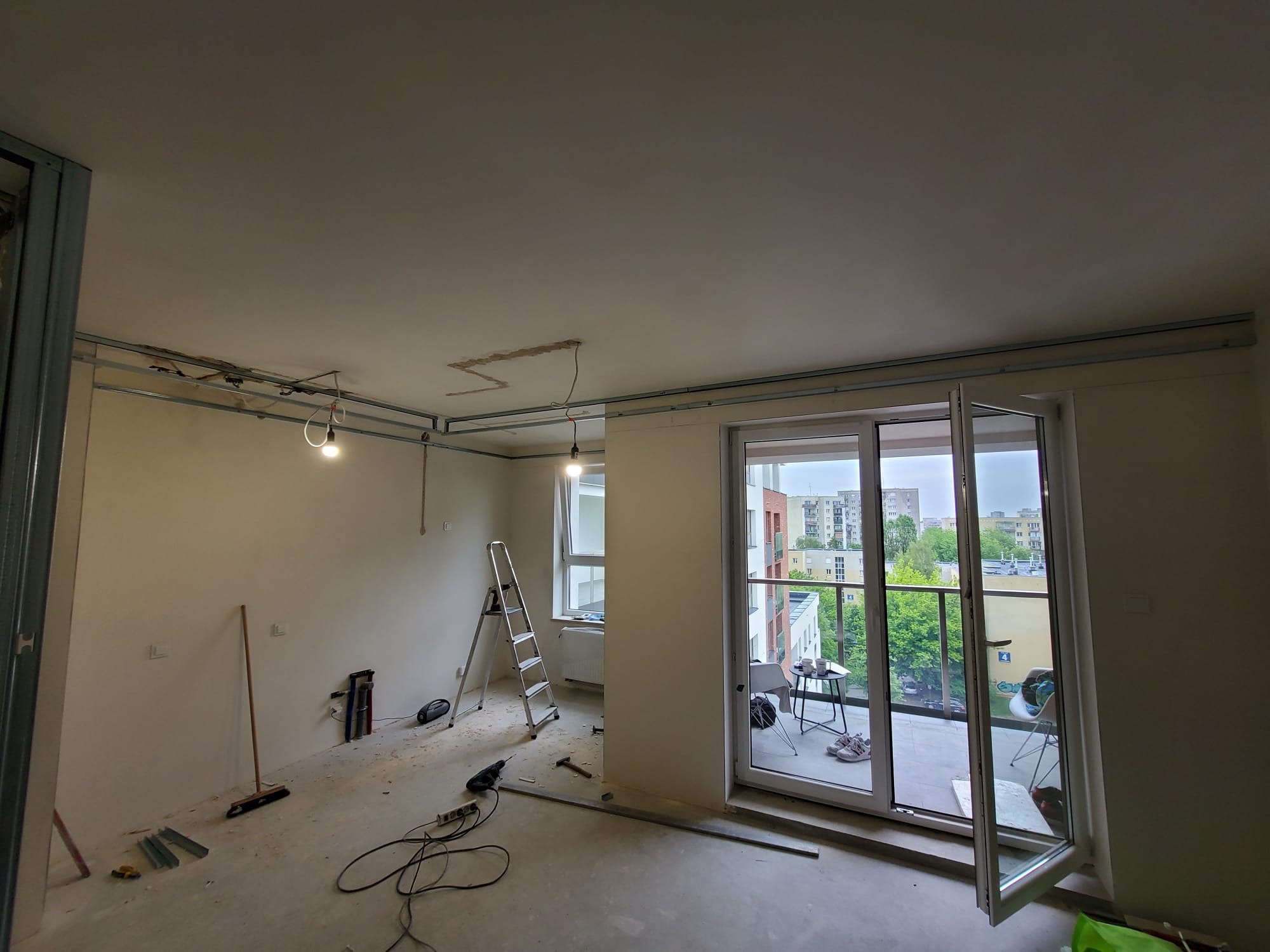 Wnętrze w trakcie remontu: widoczne profile sufitowe, drabina, narzędzia. Otwarty balkon z widokiem na miasto. Przygotowanie do montażu płyty karton-gips.
